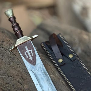 Op de afbeelding: Een decoratief zwaard met een bruin handvat en een zilveren lemmet, rustend op een houten oppervlak. Het zwaard heeft een bruine leren schede met een riem. Het lemmet heeft een symbool.