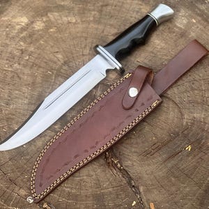 May include: A large hunting knife with a polished steel blade and a black handle. The knife is paired with a brown leather sheath with decorative stitching. The knife and sheath are displayed on a wooden surface.