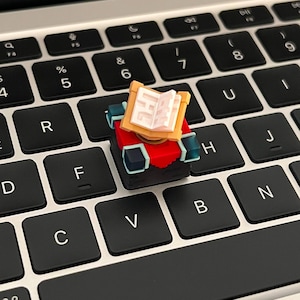 Pode incluir: Uma tecla de teclado mecânico personalizada com um livro aberto tridimensional em miniatura com capas castanho-claras e páginas brancas, assente numa base de blocos vermelha e preta com pequenos detalhes em azul-petróleo.