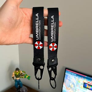 Op de afbeelding: Twee zwarte sleutelkoorden met de tekst "UMBRELLA CORPORATION" in het wit en een rood-wit paraplu-logo. Elk koord heeft een zwarte metalen sluiting en een zwarte leren lus.
