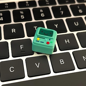 May include: A mint green custom mechanical keyboard keycap shaped like a small video game console character. The keycap features a screen with a smiling face, a yellow directional pad, a red circular button, and small blue accent buttons.