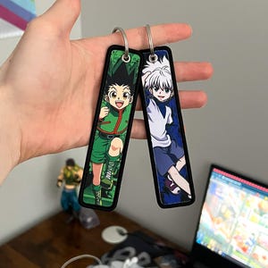 May include: Two rectangular keychains with metal rings. One keychain features a cartoon character with green hair and clothing, the other has a character with white hair and blue clothing. Both keychains have a black border.