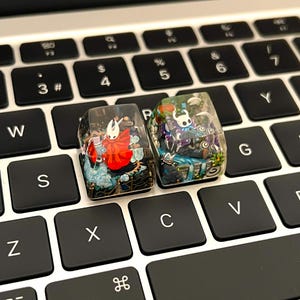May include: Two artisan mechanical keyboard keycaps made of clear resin. The left keycap features a white-masked character in a red cloak. The right keycap displays a white-masked character with blue accents, surrounded by intricate miniature landscape details.