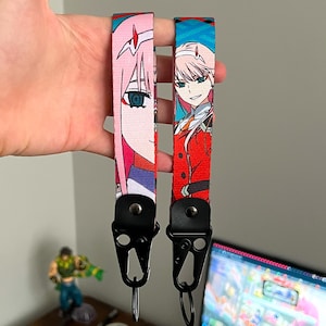 Pode incluir: Duas cordas de anime com um personagem de cabelo rosa e detalhes em vermelho. As cordas têm fechos de metal preto. O fundo está desfocado.