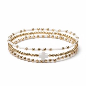 May include: A set of three beaded bracelets. The bracelets feature gold-colored beads and white beads, with a white cross charm on one bracelet. The bracelets are stacked together.