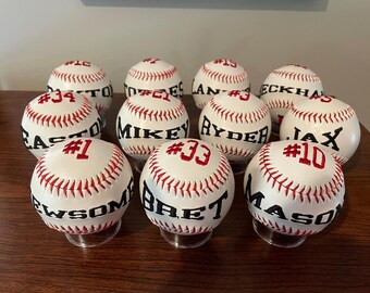 May include: A collection of personalized baseballs with red stitching and black lettering. Each baseball features a name and number, displayed on clear stands. The baseballs are white with red stitching.