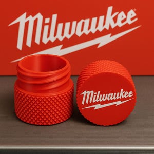 Puede incluir: Accesorio de herramienta rojo brillante con el logotipo de Milwaukee. El conjunto incluye una base cilíndrica con un patrón texturizado y una tapa a juego. El fondo presenta el logotipo de Milwaukee en blanco sobre un fondo rojo.
