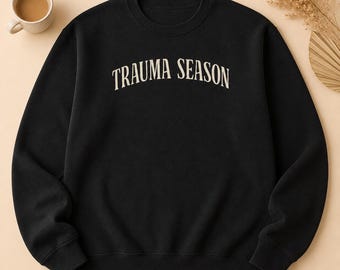 Puede incluir: Una sudadera negra de cuello redondo con el texto TRAUMA SEASON impreso en una fuente arqueada de color crema en el pecho. La prenda tiene mangas largas con puños acanalados y un dobladillo acanalado, ofreciendo un ajuste estándar.
