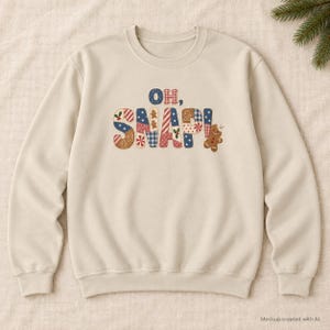 May include: A beige crewneck sweatshirt featuring the text OH, SNAP! in a patchwork-style font. The letters are filled with various patterns including blue gingham, red stripes, and gingerbread men, with a small gingerbread cookie graphic.