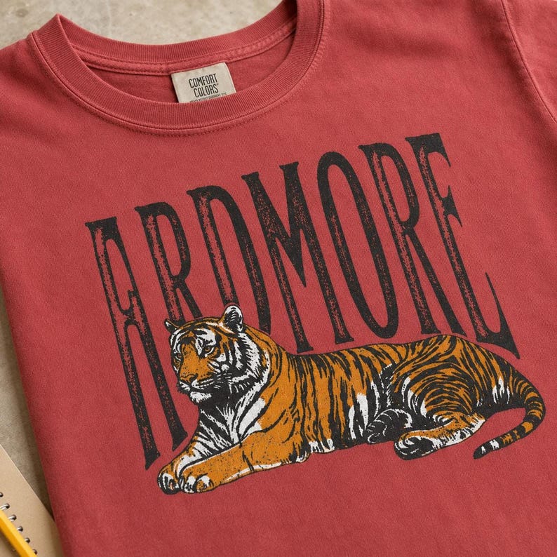 A close-up of a red t-shirt showing "ARDMORE" in black distressed letters above a lying tiger graphic, with a yellow pencil and notebook visible.