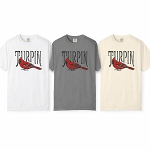 Three short-sleeve t-shirts laid flat against a white background, showing the cardinal design in white, gray, and off-white color options.