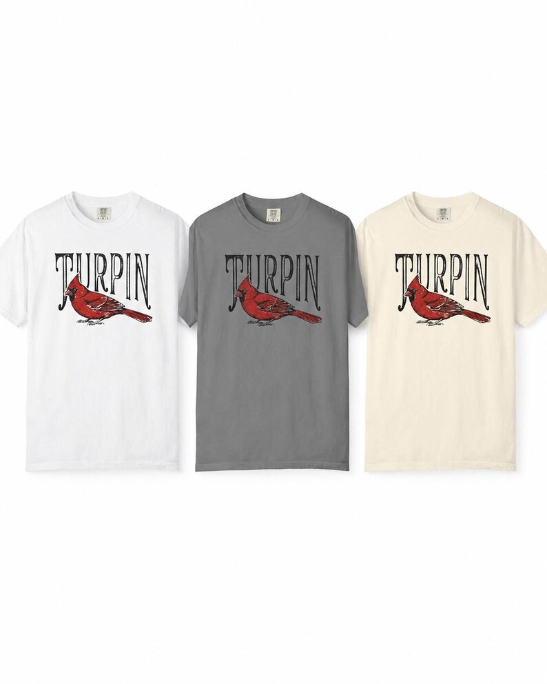 Three short-sleeve t-shirts laid flat against a white background, showing the cardinal design in white, gray, and off-white color options.