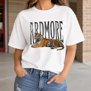 A person wearing a white t-shirt with "ARDMORE" in black distressed letters above a lying tiger graphic, with hands in jeans pockets.