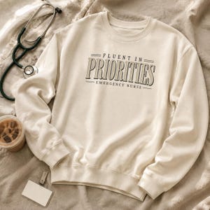 May include: A cream-colored crewneck sweatshirt featuring the text "FLUENT IN PRIORITIES" in a dark serif font, with "EMERGENCY NURSE" printed below. The design includes horizontal lines above and below the text, centered on the chest.