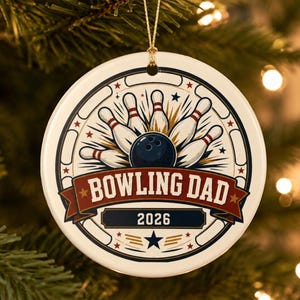 May include: A round white ceramic ornament featuring a graphic of a dark blue bowling ball hitting white pins. A red banner across the center displays the text BOWLING DAD in white letters, with the year 2026 printed in a dark blue rectangle below.