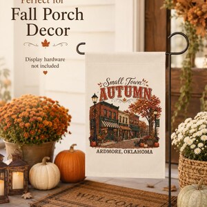 May include: A rectangular garden flag featuring a printed illustration of a small town street scene during autumn. The text reads Small Town AUTUMN and ARDMORE, OKLAHOMA. The flag is displayed on a black metal stand, showcasing a warm colour palette.
