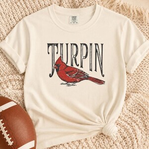 An off-white short-sleeve t-shirt with a red cardinal graphic and the word Turpin in black vintage lettering, displayed on a knitted textured surface alongside a brown football.