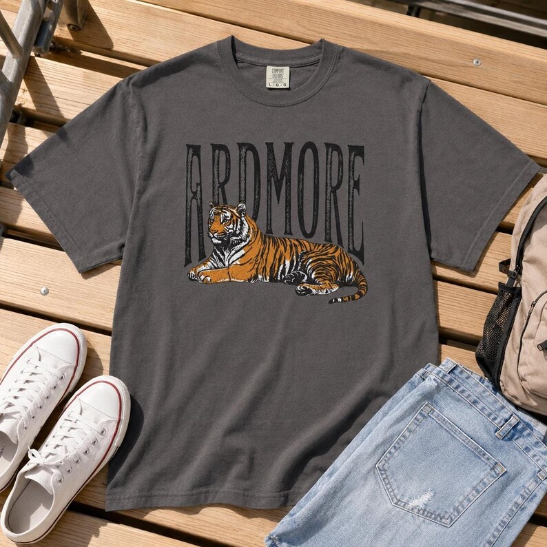 A dark gray t-shirt with "ARDMORE" in black distressed letters above a lying tiger graphic, displayed on a wooden surface with white sneakers and jeans.