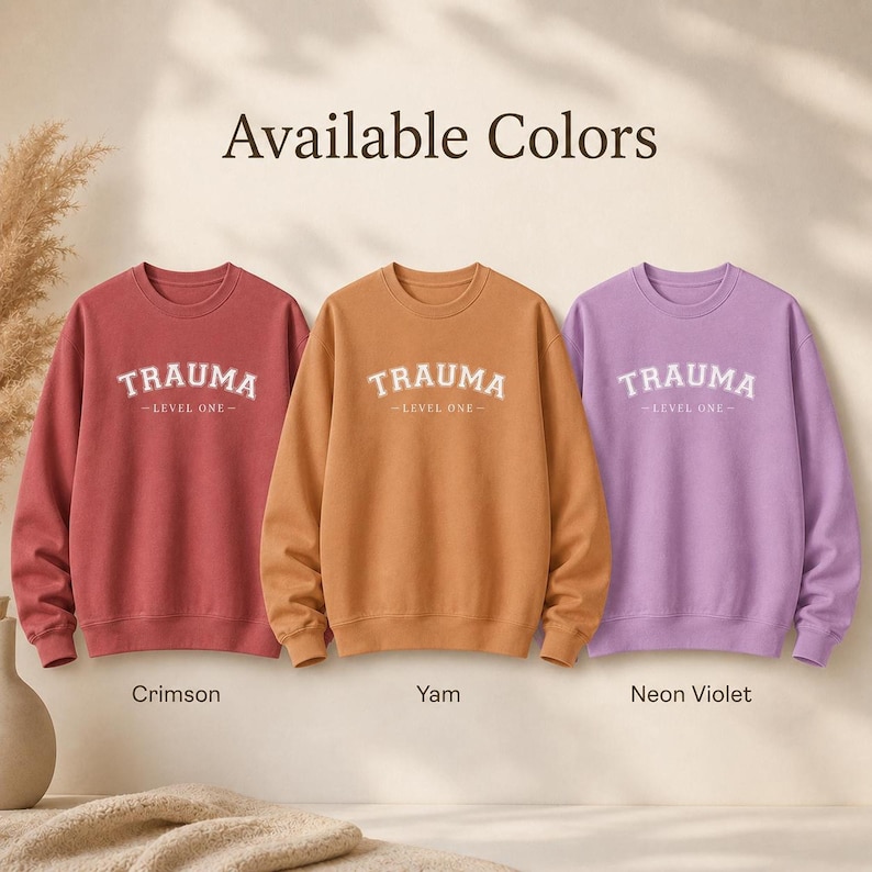 Three crewneck sweatshirts in Crimson, Yam, and Neon Violet, each featuring the TRAUMA LEVEL ONE print.