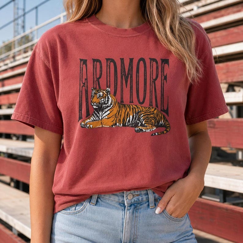 A person wearing a red t-shirt with "ARDMORE" in black distressed letters above a lying tiger graphic, standing outdoors with stadium bleachers in the background.
