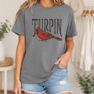 A woman wearing the gray cardinal graphic t-shirt and distressed denim shorts, standing indoors with clothing racks and greenery in the background.