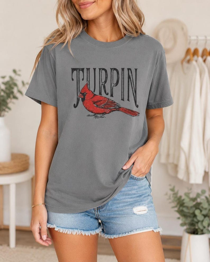 A woman wearing the gray cardinal graphic t-shirt and distressed denim shorts, standing indoors with clothing racks and greenery in the background.