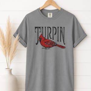 A gray short-sleeve t-shirt with the cardinal and Turpin text hanging on a wooden hanger against a white paneled wall, next to a white vase with dried grass.