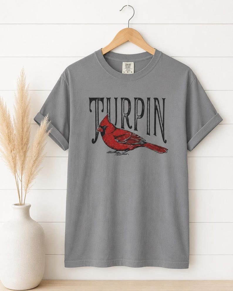 A gray short-sleeve t-shirt with the cardinal and Turpin text hanging on a wooden hanger against a white paneled wall, next to a white vase with dried grass.