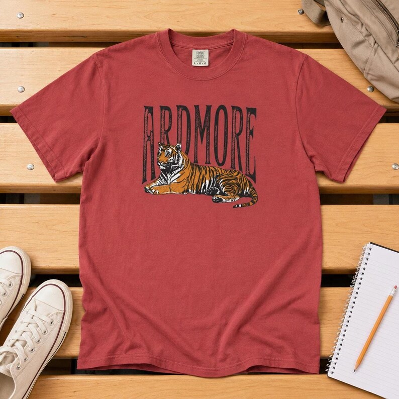 A red t-shirt with "ARDMORE" in black distressed letters above a lying tiger graphic, displayed on a wooden surface with white sneakers and a backpack.