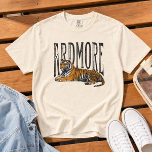 A cream t-shirt with "ARDMORE" in black distressed letters above a lying tiger graphic, displayed on a wooden surface with white sneakers and a denim jacket.