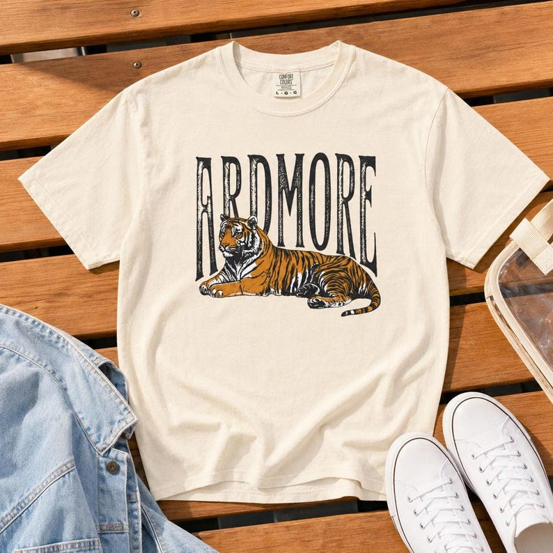 A cream t-shirt with "ARDMORE" in black distressed letters above a lying tiger graphic, displayed on a wooden surface with white sneakers and a denim jacket.