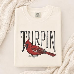 A neatly folded cream-colored t-shirt with the red cardinal graphic and Turpin lettering, resting on a white knitted blanket.