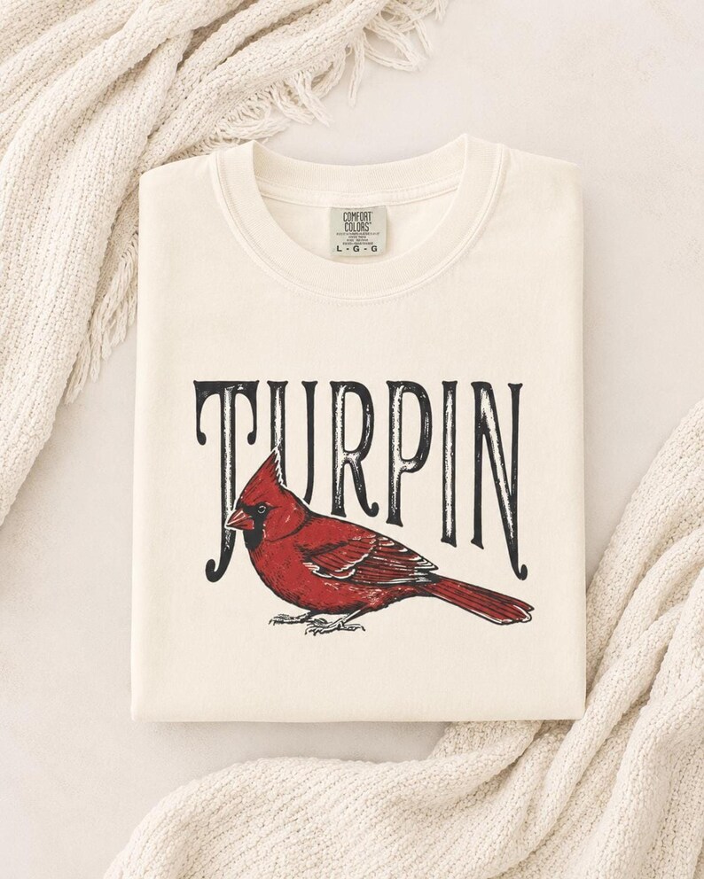 A neatly folded cream-colored t-shirt with the red cardinal graphic and Turpin lettering, resting on a white knitted blanket.