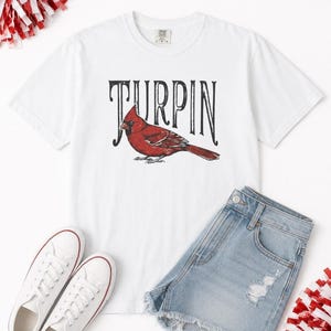 Flat lay of a white cardinal graphic t-shirt paired with light blue denim shorts, white sneakers, and red and white pom-poms.
