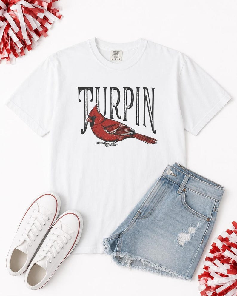 Flat lay of a white cardinal graphic t-shirt paired with light blue denim shorts, white sneakers, and red and white pom-poms.