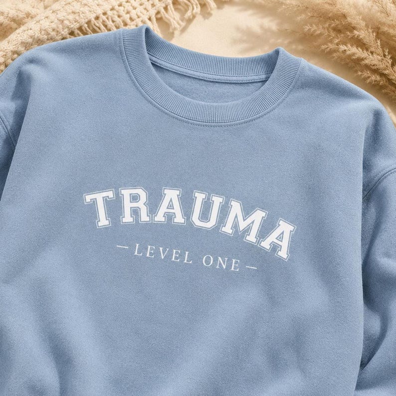 Close-up of the light blue sweatshirt fabric showing the white TRAUMA LEVEL ONE lettering.