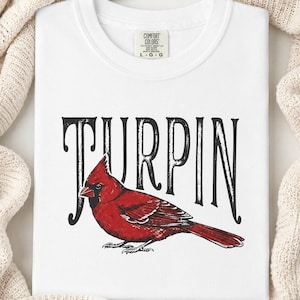 A close-up of a folded white t-shirt displaying a detailed red cardinal graphic and black Turpin lettering, placed on a textured knit fabric.