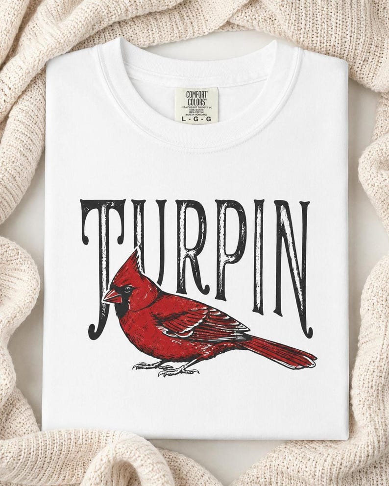 A close-up of a folded white t-shirt displaying a detailed red cardinal graphic and black Turpin lettering, placed on a textured knit fabric.
