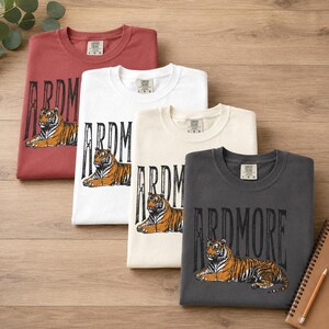 Four t-shirts in red, white, cream, and dark gray, each with "ARDMORE" and a lying tiger graphic, laid out diagonally on a wooden surface.
