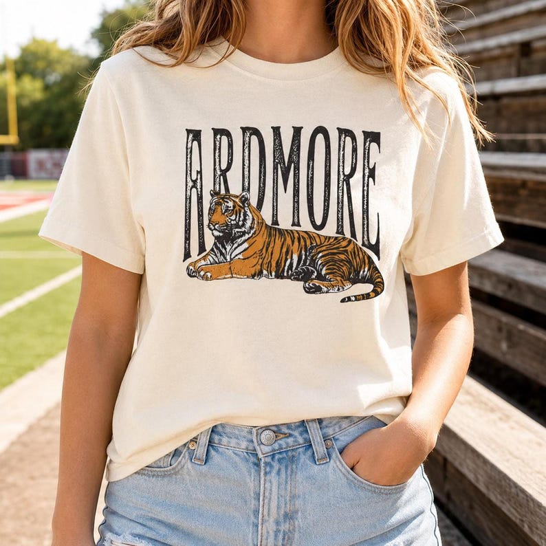 A person wearing a cream t-shirt with "ARDMORE" in black distressed letters above a lying tiger graphic, standing outdoors with stadium bleachers in the background.