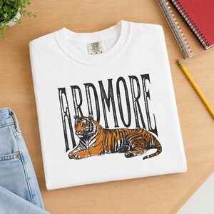A white t-shirt with "ARDMORE" in black distressed letters above a lying tiger graphic, displayed on a wooden surface next to a notebook and pencil.
