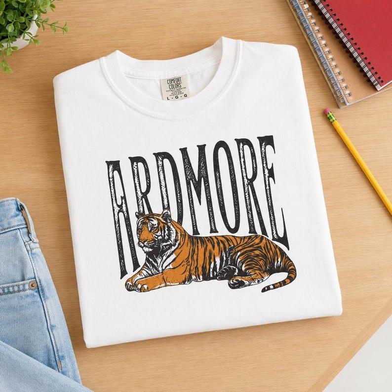 A white t-shirt with "ARDMORE" in black distressed letters above a lying tiger graphic, displayed on a wooden surface next to a notebook and pencil.
