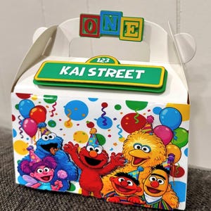 May include: A white cardboard party favor box featuring a colorful confetti pattern and Sesame Street characters including Elmo, Big Bird, Cookie Monster, Abby Cadabby, Bert, and Ernie. The top displays a green street sign reading KAI STREET.