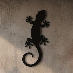 Puede incluir: Una cama de metal negro con una colcha y almohadas grises. Una silueta de lagarto negro está montada en la pared sobre la cama. Dos lámparas colgantes con bombillas Edison flanquean la cama.