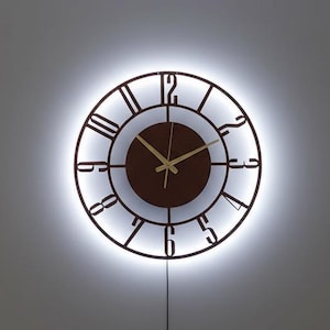 May include: A modern, round wall clock with a brown face and gold hands. The clock features Roman numerals and is surrounded by a glowing white light. The clock is mounted on a gray wall.