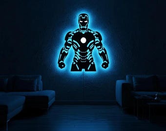 May include: A wall-mounted LED light fitting shaped like the silhouette of a superhero in armour. The figure is black with intricate cut-out details, illuminated from behind by a bright blue glow that outlines the entire form and a white circle.