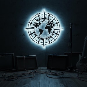 Pode incluir: Uma peça de arte de parede circular e brilhante com um mapa-múndi dentro de um design de rosa dos ventos. A peça é iluminada com uma luz azul fria, montada em uma parede escura. Amplificadores, um microfone e uma guitarra na sala.
