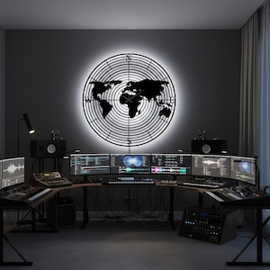 Puede incluir: Un reloj negro con forma de mapa del mundo está montado en una pared gris oscura. El reloj está rodeado por una luz brillante. Debajo hay una estación de trabajo con múltiples monitores de computadora, teclados y equipos de audio.