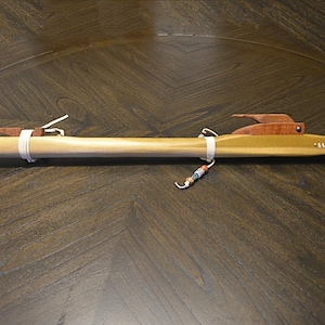 May include: A handcrafted wooden flute with a natural finish. The flute features decorative elements, including leather accents and a carved wooden bird design. The instrument is secured with white bands and adorned with beaded details. The flute is displayed on a dark wooden surface.