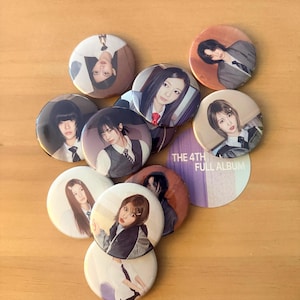 May include: A collection of round buttons featuring images of people in various poses and outfits. Some buttons have the text "THE 4TH FULL ALBUM" printed on them. The buttons are arranged on a light brown surface.
